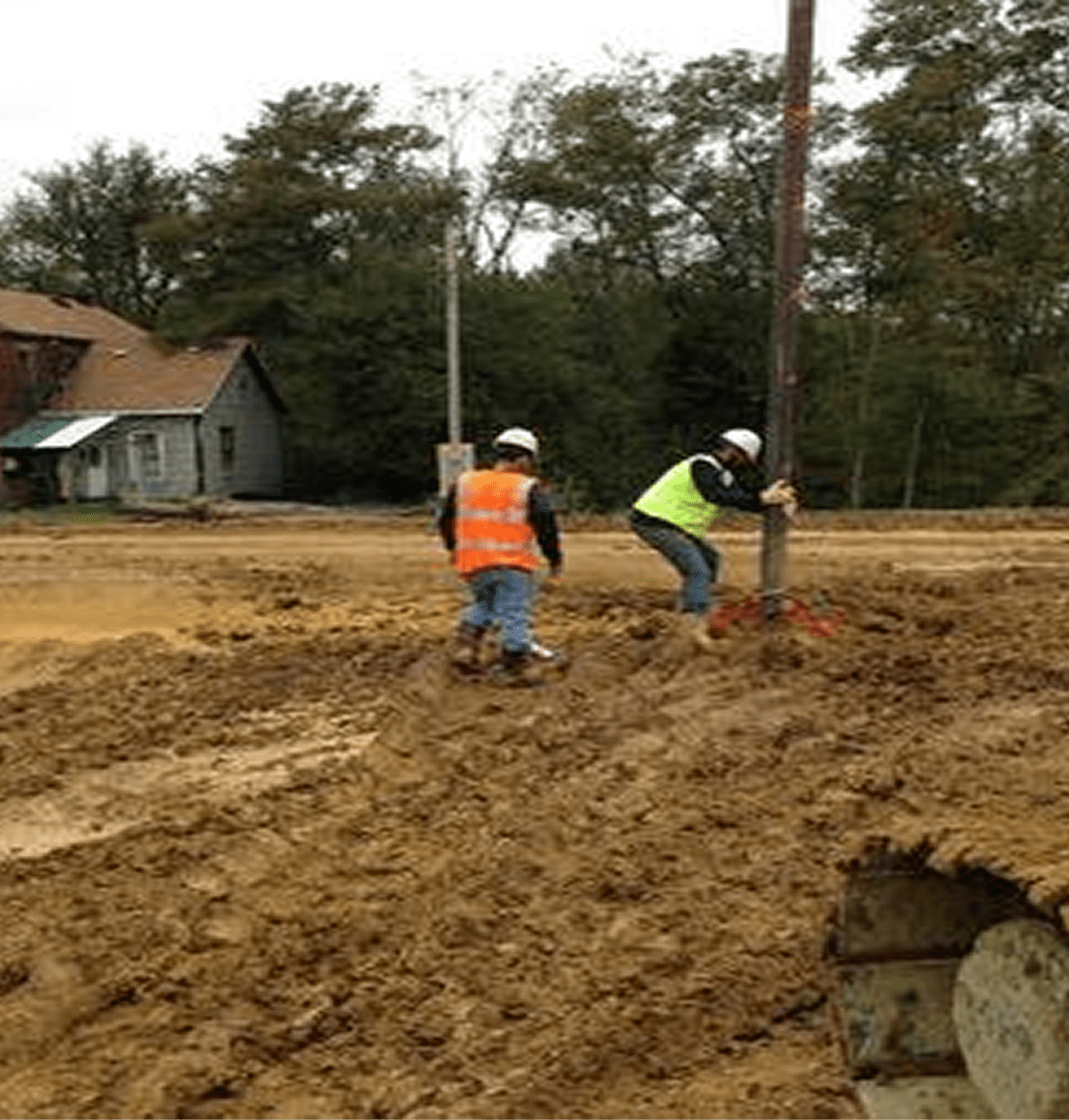 Two construction workers working on a muddy site near houses.