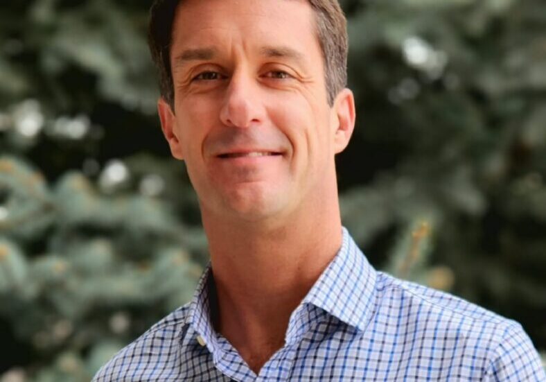 Smiling man in a checkered shirt outdoors with blurred greenery background.