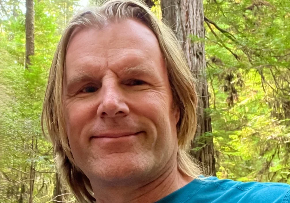 Smiling man in a blue shirt outdoors with trees in the background.